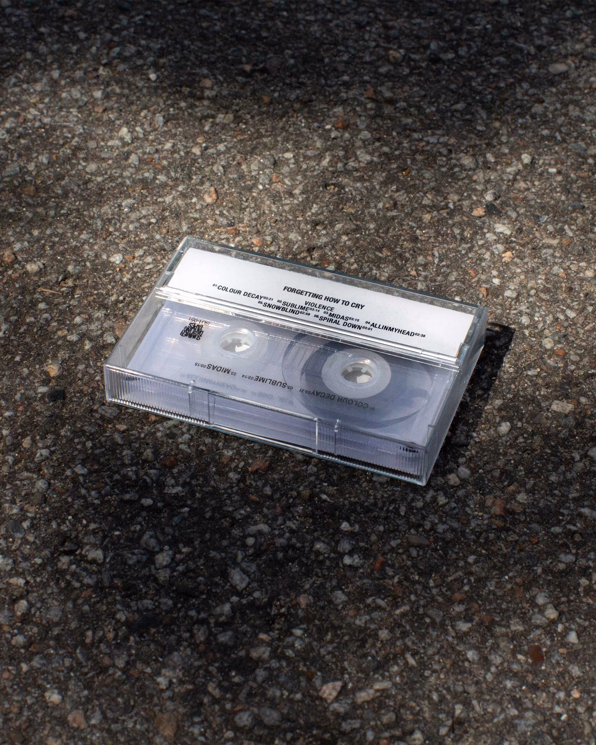 Clear cassette tape in its case resting on a textured asphalt surface under sunlight and shadows.