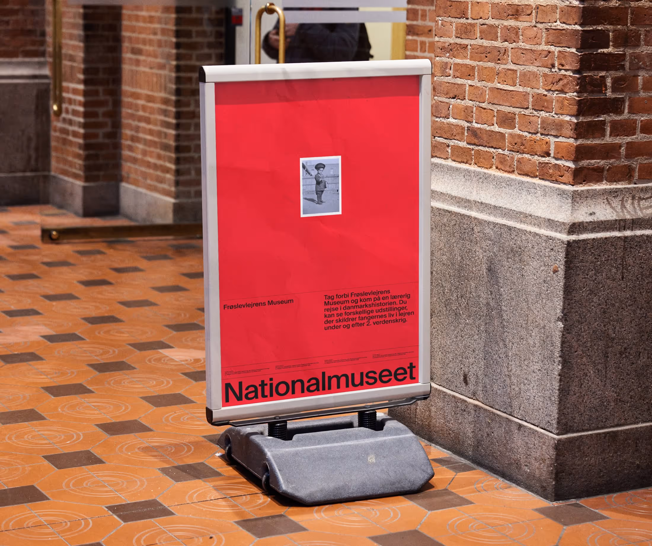 Red Nationalmuseet signboard with a small black-and-white photo of a person in uniform, placed on a patterned tile floor beside a brick wall.
